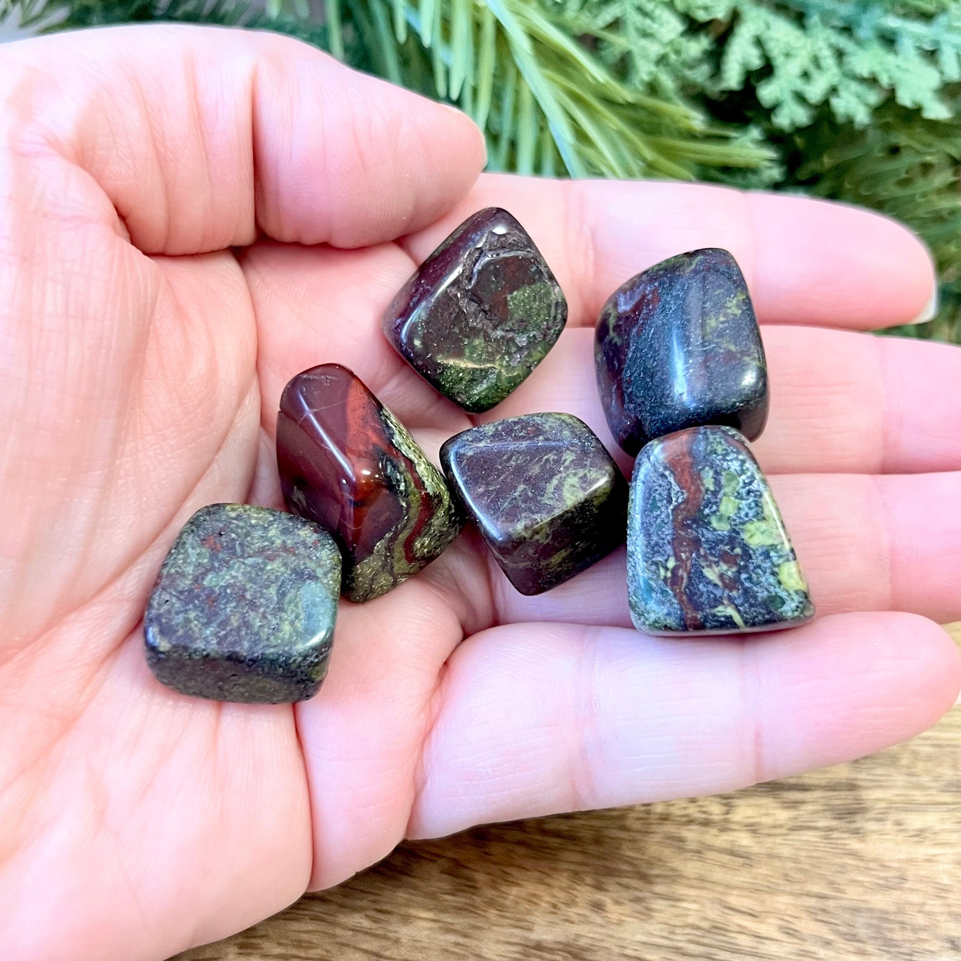 Group of green and red tumbled crystals in a person's hand. They are Dragon Blood Stone, and a polished cube shape.
