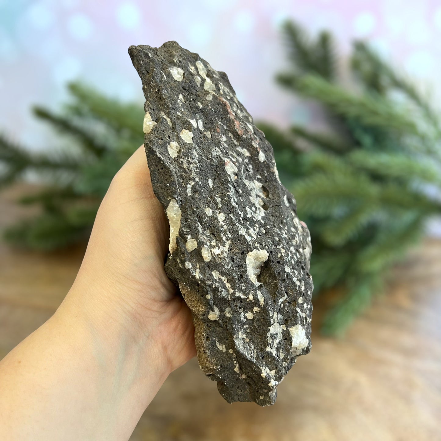 Image of a large, rough chunk of black Lava Rock Stone on a wooden table. Has rough texture with lots of holes, and white Quartz growing in spots.