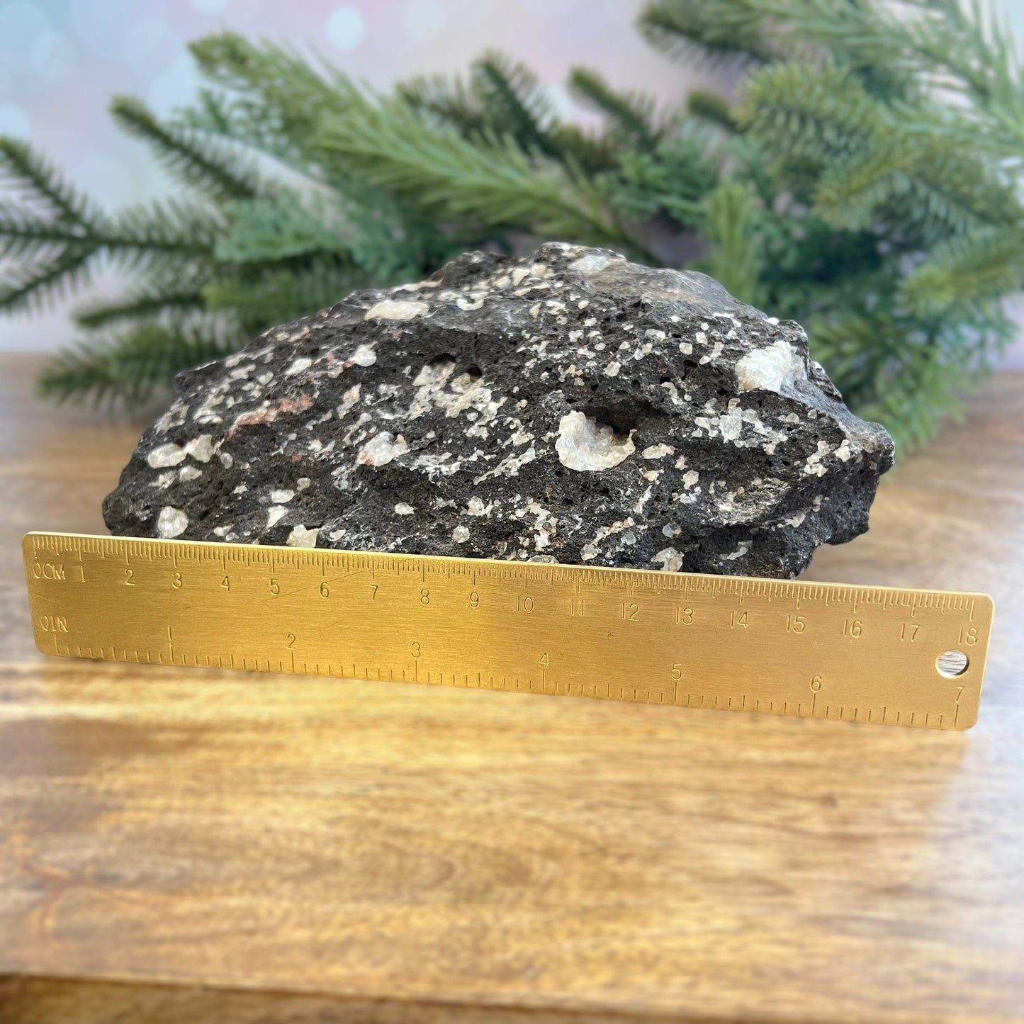 Image of a large, rough chunk of black Lava Rock Stone on a wooden table. Has rough texture with lots of holes, and white Quartz growing in spots.