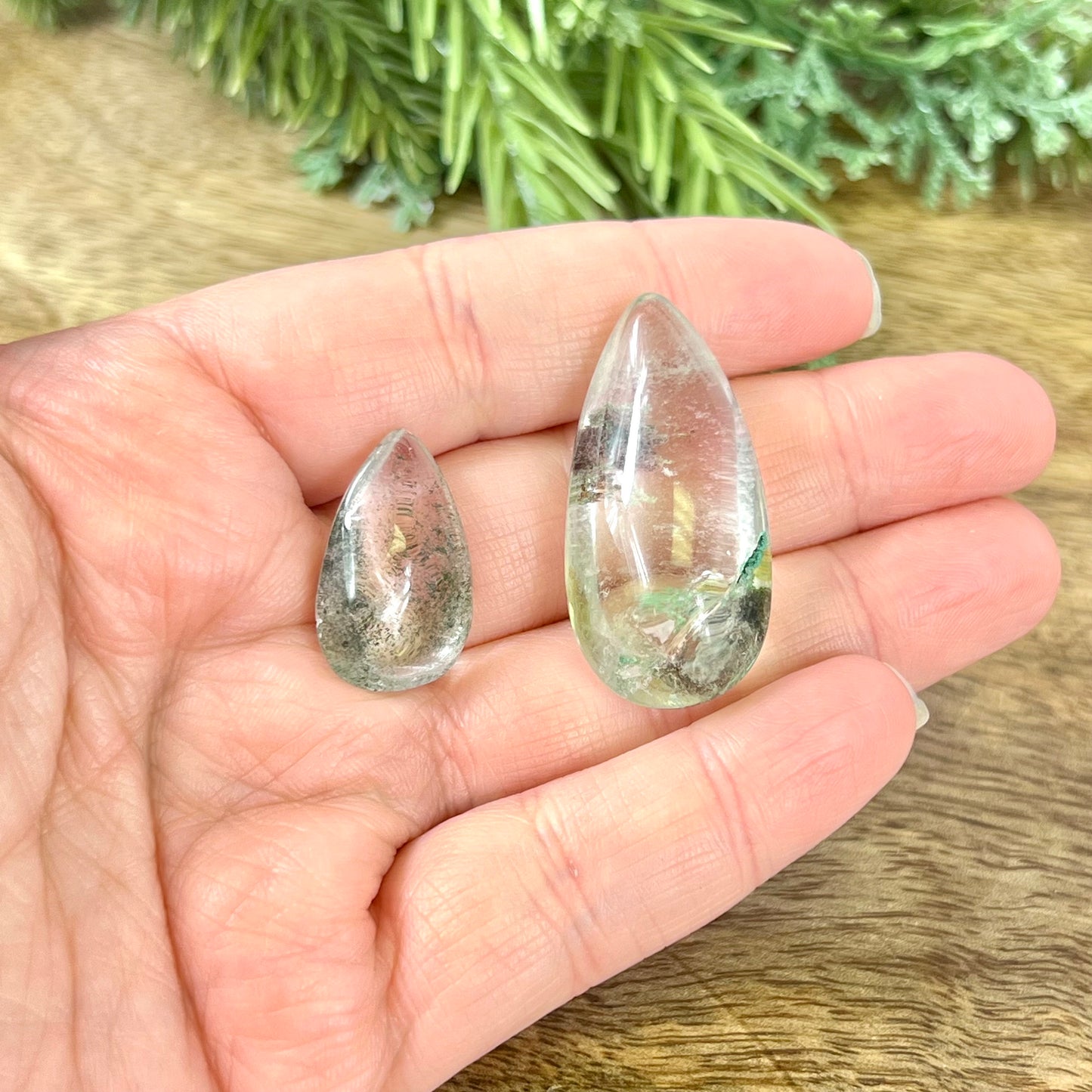 A person's hand holding a group of crystals polished into a teardrop shape. They're a clear quartz with green Chlorite inclusions.