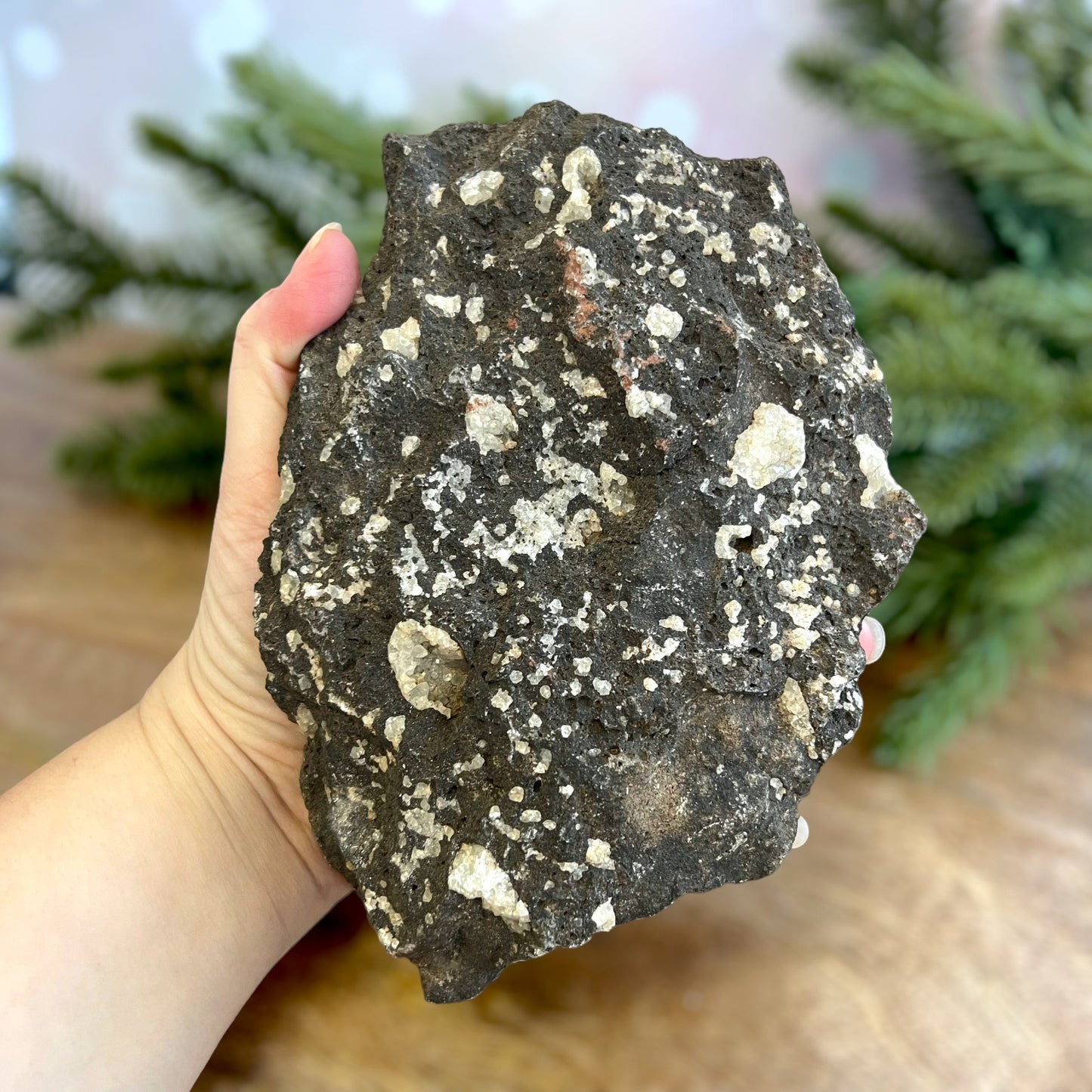 Image of a large, rough chunk of black Lava Rock Stone on a wooden table. Has rough texture with lots of holes, and white Quartz growing in spots.