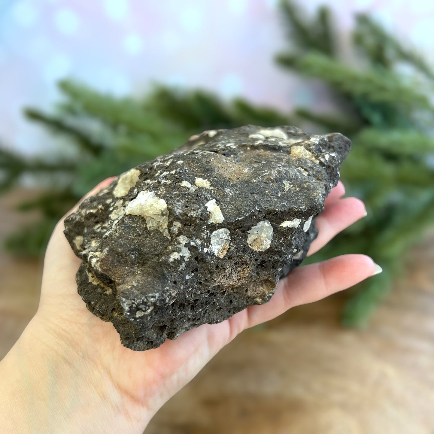 Image of a large, rough chunk of black Lava Rock Stone on a wooden table. Has rough texture with lots of holes, and white Quartz growing in spots.