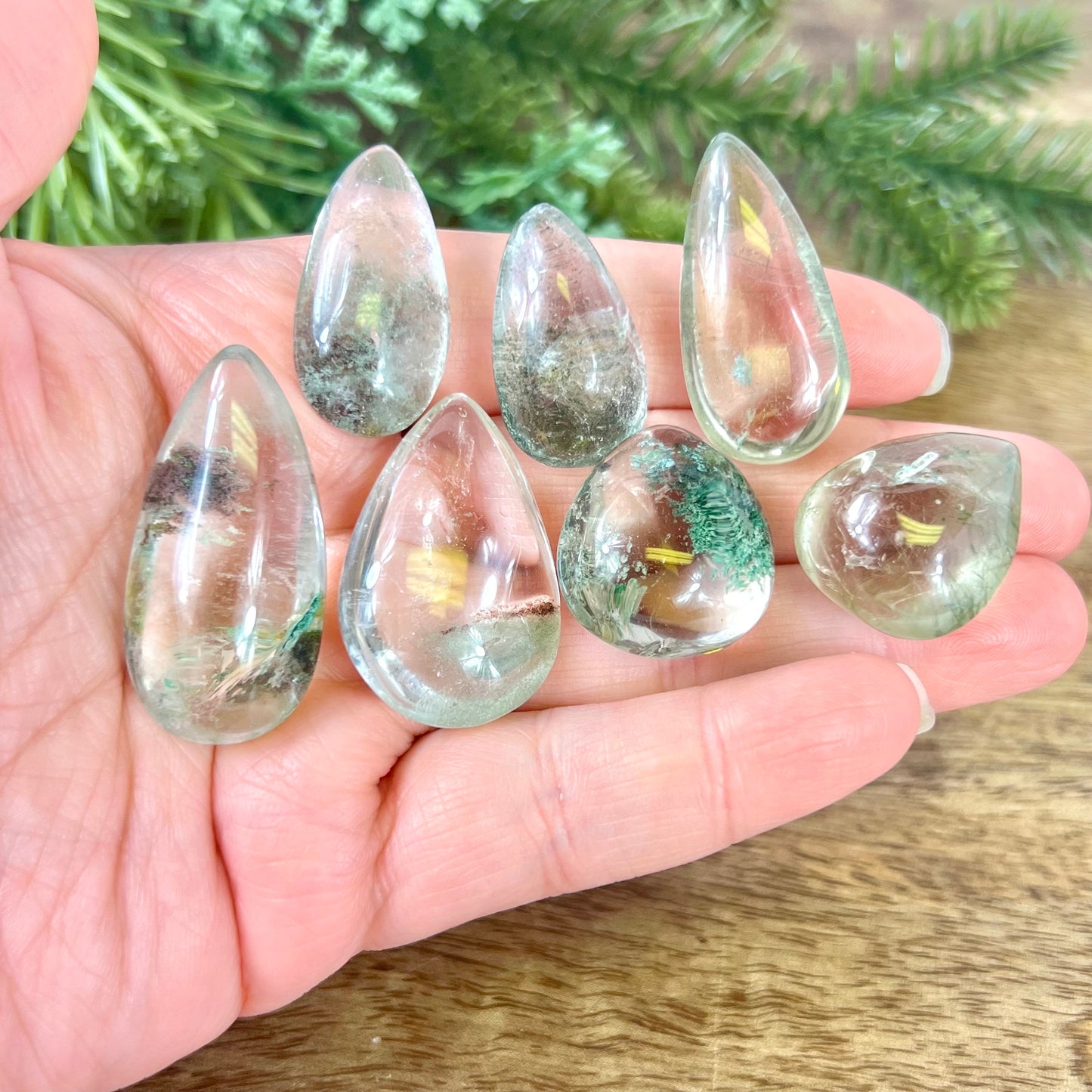 A person's hand holding a group of crystals polished into a teardrop shape. They're a clear quartz with green Chlorite inclusions.