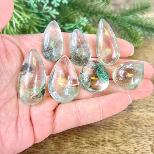 A person's hand holding a group of crystals polished into a teardrop shape. They're a clear quartz with green Chlorite inclusions.