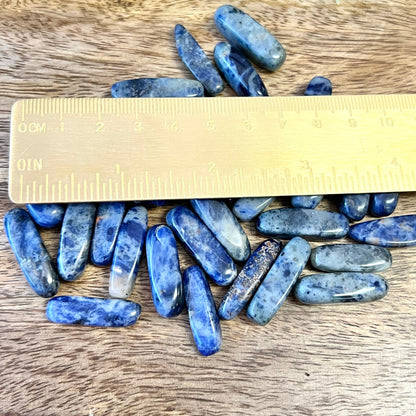 a group of blue and white sodalite tumbled crystals next to a gold ruler. They are long oval dagger shapes