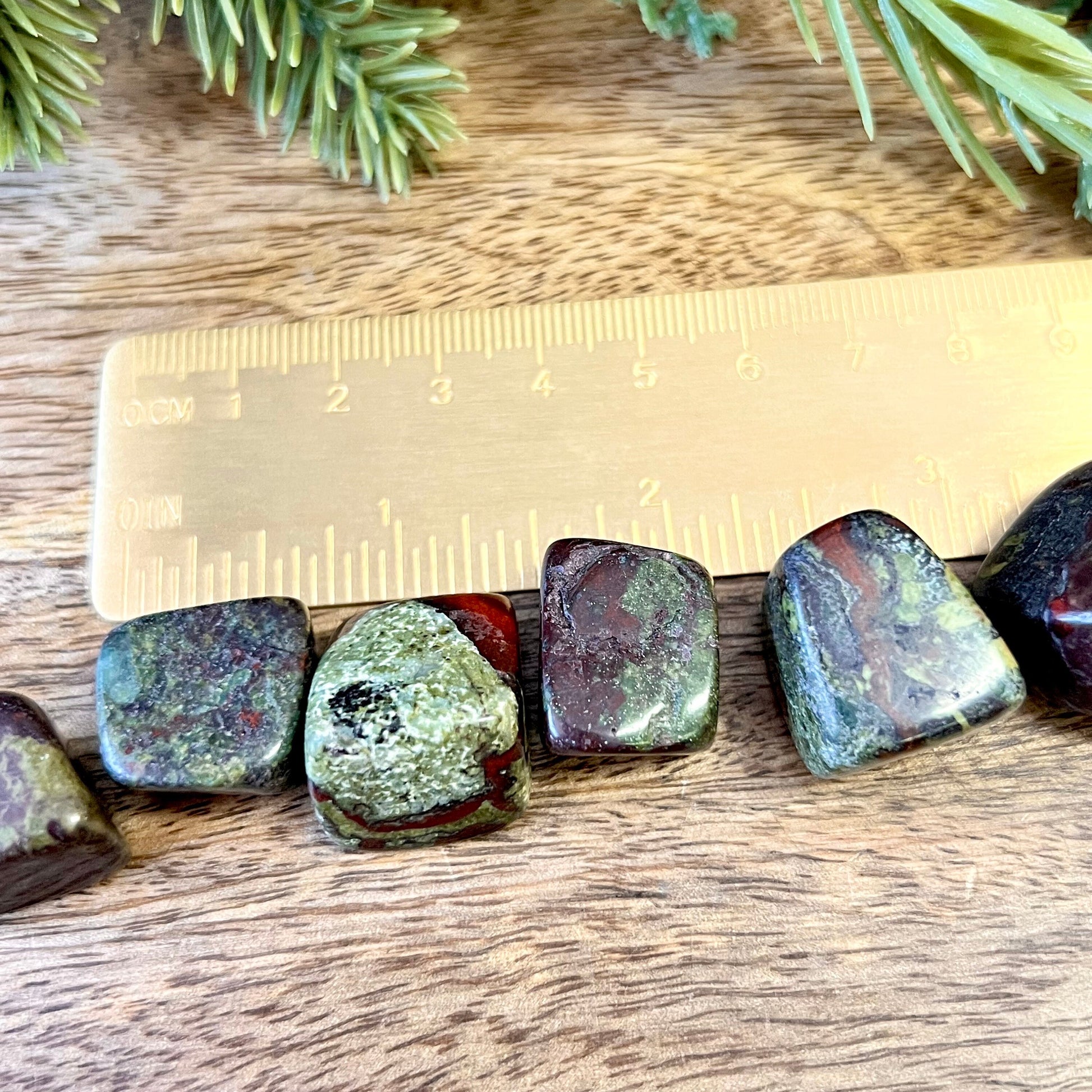 Group of green and red tumbled crystals next to a ruler. They are Dragon Blood Stone, and a polished cube shape.