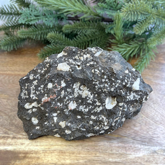 Image of a large, rough chunk of black Lava Rock Stone on a wooden table. Has rough texture with lots of holes, and white Quartz growing in spots.