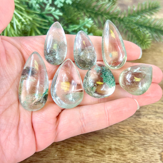 A person&#39;s hand holding a group of crystals polished into a teardrop shape. They&#39;re a clear quartz with green Chlorite inclusions.