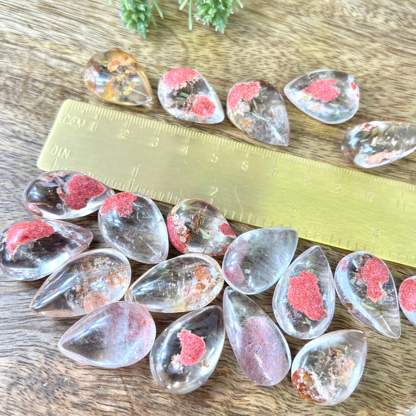 a group of dyed red Garden Quartz tumbled crystals in a teardrop shape next to a gold ruler